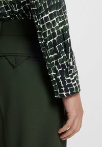 Camicia a maniche lunghe con un motivo geometrico verde e bianco, abbinata a pantaloni verdi a vita alta con dettaglio dei bottoni.
