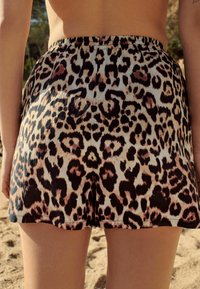 Personne portant un short en imprimé léopard se tenant à l'extérieur sur un sol sablonneux avec des arbres en arrière-plan.