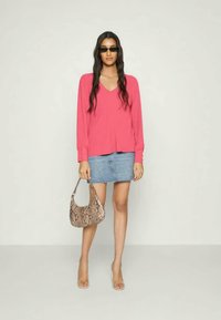 Top rosa de mangas longas em V, saia mini em denim azul claro, e uma bolsa de mão em estampa de cobra bege. Modelo usa óculos de sol pretos e saltos transparentes.