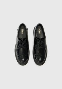 Musta nahkainen brogue-kengät kiiltävällä pinnalla, pyöreällä kärjellä, reikäkoristeilla ja mustalla kumipohjalla. Brändin nimi "Clarks" pohjallisissa.