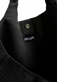 Svart textil toteväska med en rynkad struktur, med en knappstängning och en synlig etikett med texten "PIECES" på insidan.