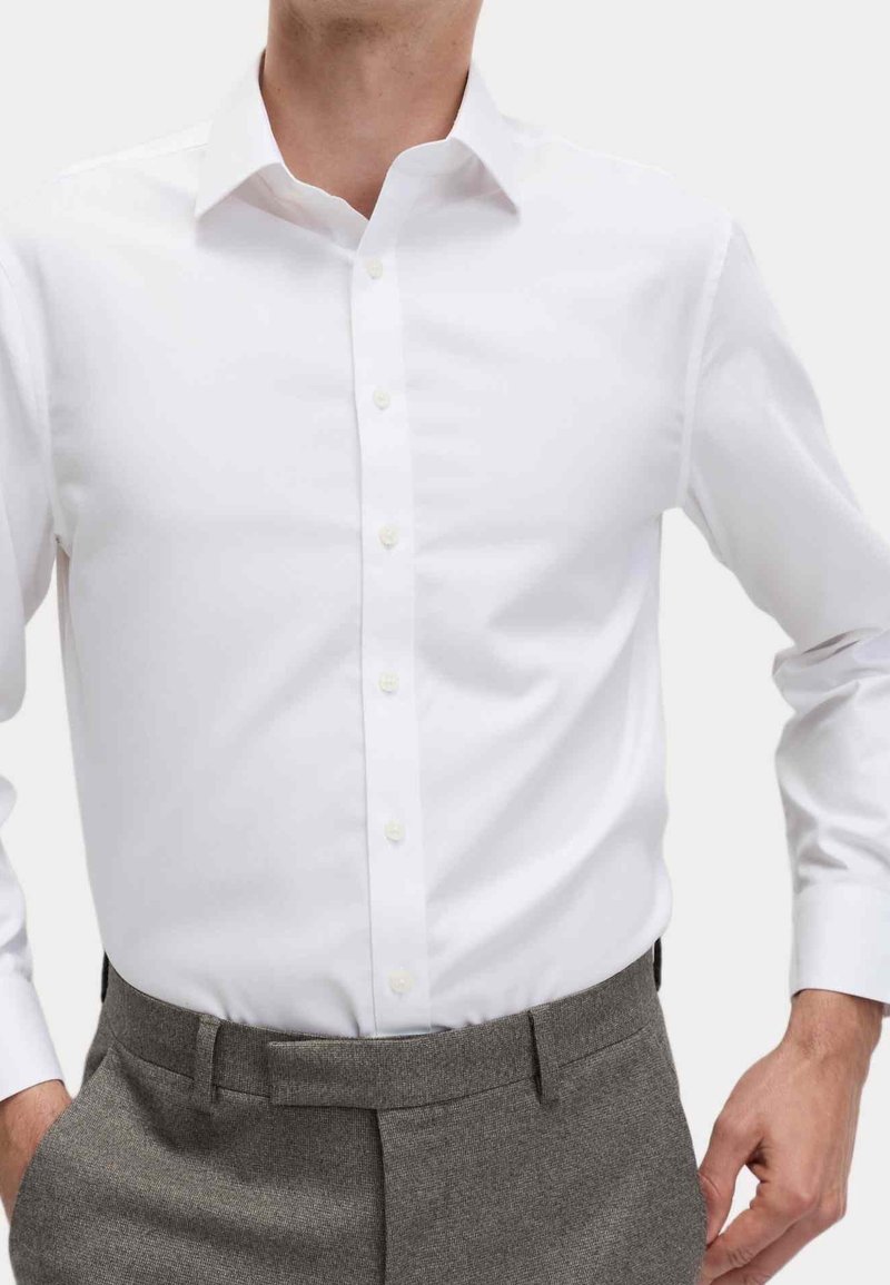 Homme portant une chemise blanche à manches longues rentrée dans un pantalon gris, les mains près des poches.
