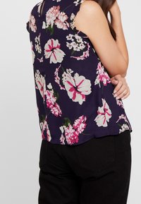 Květinová blůza s námořnickým pozadím, zdobená velkými růžovými a bílými květy, bez rukávů a s vlnitým detailem na ramínkách.