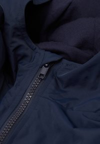 Vnitřek navy modré bundy se hladkým textilním povrchem, kontrastním tmavým zipem a měkkou, texturovanou podšívkou viditelnou.