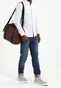 Bolso mensajero de cuero marrón con correas negras, llevado sobre una camisa blanca de botones, jeans azules y zapatillas altas grises.