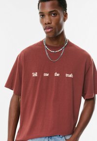 T-shirt en coton maroon à manches courtes, présentant le texte "Dis-moi la vérité" en lettres rose pâle sur le devant. Coupe décontractée.