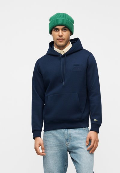 Homme portant un bonnet vert, un sweat à capuche bleu marine avec poche avant, une chemise beige en dessous, et un jean bleu clair, debout devant un fond uni.