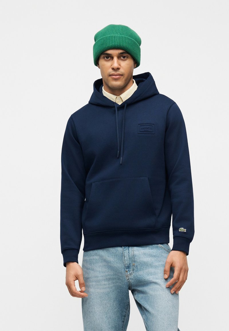 Άνδρας που φοράει πράσινο σκουφάκι, μπλε-ναυτικό φούτερ με μπροστινή τσέπη, μπεζ πουκάμισο από κάτω και ανοιχτόχρωμο μπλε τζιν, στέκεται μπροστά σε απλή φόντο.