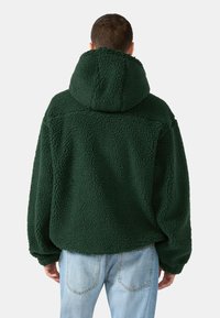 Zelená fleecová bunda s textúrovanou, nadýchanou tkaninou, má kapucňu a voľný strih. V kombinácii so svetlomodrými džínsami. Pohľad zozadu.