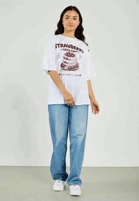 Noor naine seisab, seljas valge ülemõõduline T-särk, millel on maasikakooki kujutav graafika, ning sinised teksad, lihtsa heleda tausta ees.