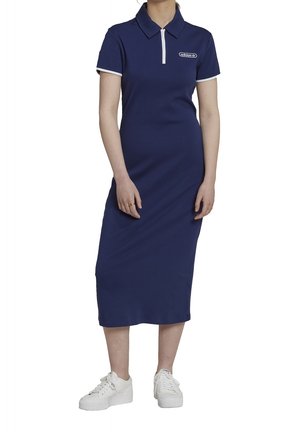 Robe polo côtelée bleu marine avec col blanc et fermeture éclair, arborant un logo brodé sur la poitrine et associée à des baskets blanches.