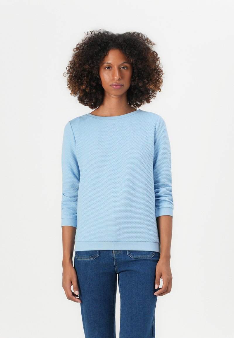 Haut long à manches longues bleu clair texturé avec un col rond et des poignets côtelés, associé à un jean bleu foncé. Design simple et moderne.