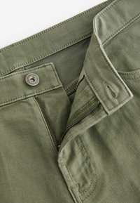 Pantalons en coton verts avec zip et fermeture à bouton métallique. Texture lisse avec coutures renforcées et accents cousus.