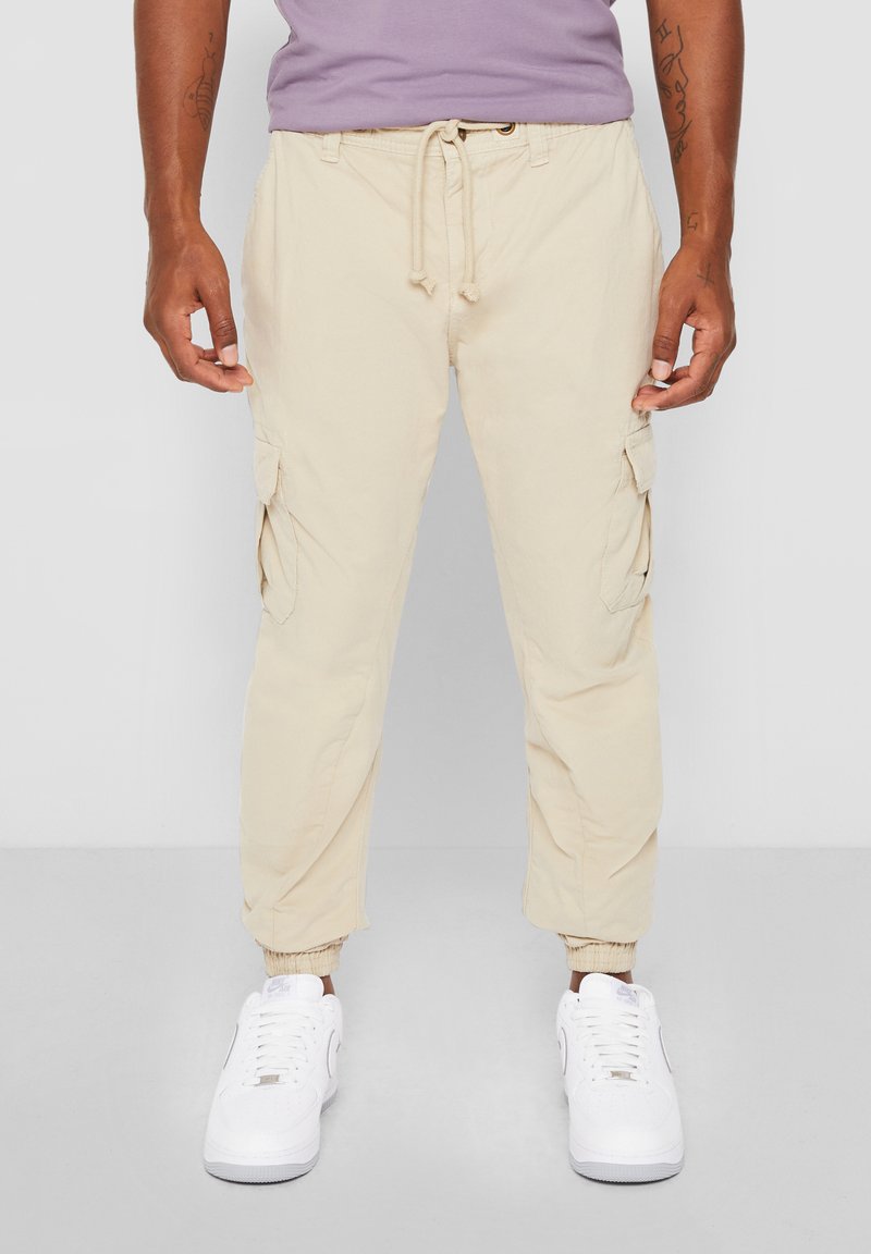 Hombre con pantalones jogger cargo beige con cintura ajustable y puños elásticos, combinado con zapatillas blancas y camisa morada.