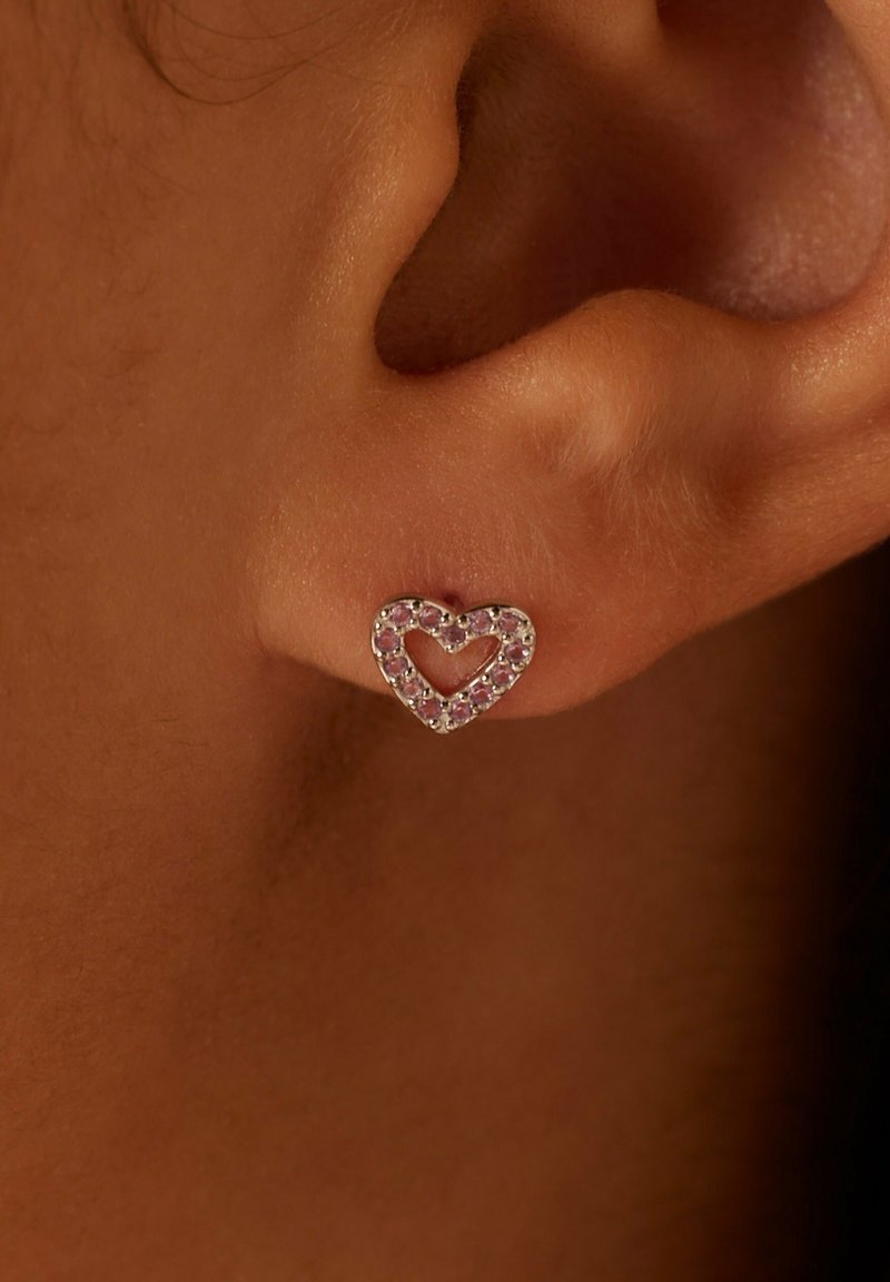 Zilveren hartvormige oorbel met roze edelsteenaccenten, met een open centrumontwerp en een gepolijste afwerking, weergegeven op een close-up van de huid.