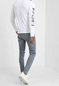 Camisa blanca de manga larga con impresiones gráficas negras en las mangas, combinada con jeans ajustados de color azul claro y zapatillas blancas. Diseño sencillo, ajuste casual.