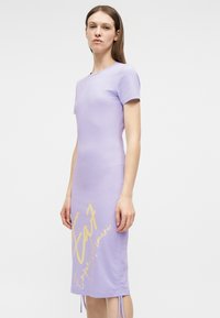 Γυναίκα φορώντας στενή λιλά φόρεμα με κοντά μανίκια και κίτρινο κείμενο «Emporio Armani» κοντά στο τελείωμα, στέκεται μπροστά σε απλό φόντο.