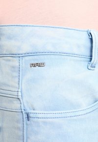Vaqueros de mezclilla azul claro con una textura desgastada, que presentan un pequeño logo plateado "RAW" en el bolsillo trasero.