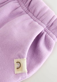 Világos lila kötött anyag, ráncolt gumidétellel és egy világosbarna anyagcímkével, amelyen a szélénél egy félkör alakú minta van hímezve.