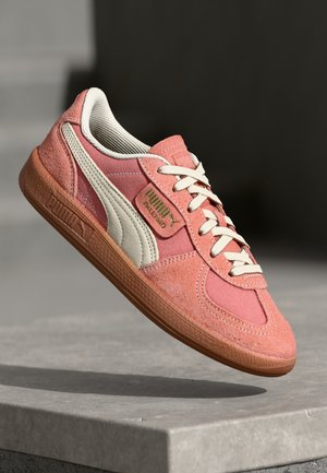 Baskets Puma roses et blanches avec semelle en gomme, lacets blancs et logo Puma, flottant au-dessus d'une surface en béton gris et d'un fond sombre.