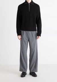 Schwarzer gerippter Strickpullover mit einem Reißverschluss-Kragen, kombiniert mit hellgrauen, weiten Hosen und schwarzen Lederschuhen.