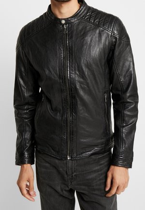 Homme portant une veste en cuir noire ajustée avec des détails matelassés aux épaules et des poches zippées à l'avant et sur les côtés, associée à un jean foncé.