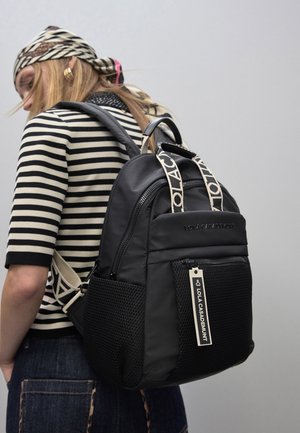 Mochila negra de nylon con bolsillo lateral de malla, detalle de logo en la cinta y compartimentos con cremallera. Superficie texturizada y características de diseño ergonómico.