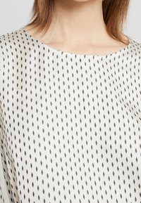 Tissu de couleur claire présentant un motif en diamant de couleur gris foncé. Le haut a un col rond et une texture lisse et douce.