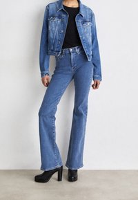 Woman wearing a cropped blue denim jacket, high-waisted blue flared jeans, black top, and black heeled boots against a plain wall.