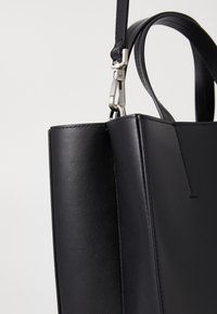 Geantă tote din piele neagră, cu textură netedă, formă rectangulară și detalii din metal argintiu. Include un mâner superior și cusături pe laturi.