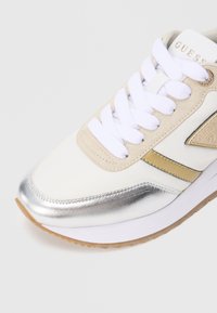 Sneaker con tomaia bianca e beige, dettagli metallici argentati, lacci bianchi e suola beige testurizzata. Presenta una finitura simile alla pelle.