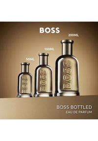 Tres botellas de cristal de Boss Bottled Eau de Parfum en tamaños de 50 ml, 100 ml y 200 ml, mostradas sobre un fondo degradado marrón.