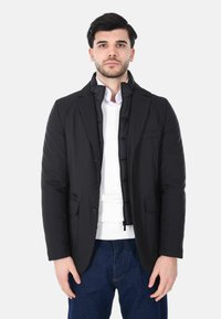 Giacca esterna nera con imbottitura interna trapuntata, rever a lancia e due tasche frontali, abbinata a un maglione bianco e jeans blu.