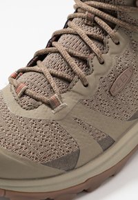 Scarpa da trekking beige con tomaia in maglia testurizzata, punta rinforzata e elementi a contrasto. Si notano lacci spessi e una suola in gomma robusta.