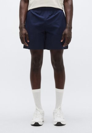 Jūras flotes šorti ar elastīgu vidukļa jostu, valkāti kopā ar baltiem sporta zeķēm un baltiem sporta apaviem. Gluda tekstūra un taisna griezuma dizains.