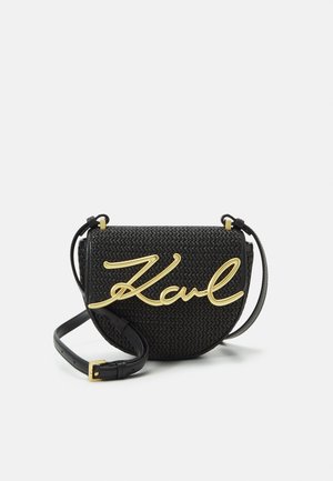 Černá tkaná crossbody taška s zakřiveným tvarem, s velkým zlatým logem "Karl" a štíhlým nastavitelným popruhem. Texturovaný povrch.