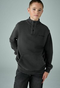 Tamsiai pilkas megztinis su aukštu apykakle, trijų mygtukų užsegimu, priekinėmis kišenėmis ir raštuotais rankogaliais. „ALLSAINTS“ logotipas išsiuvinėtas ant krūtinės.