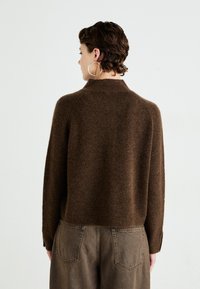 Person with short curly hair wearing large hoop earrings, a brown fuzzy sweater, and faded brown pants, facing away against a plain background.