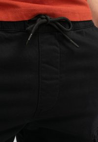 Zwarte joggers met elastische taille en een trekkoord, met een gladde textuur en subtiele stikseldetails. Oranje top zichtbaar bij de taille.