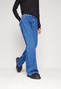 Blue denim wide-leg jeans with a high waist, featuring a classic five-pocket design and contrasting stitching. Worn with a black top and shoes.