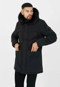 Svart parkas med konstpäls på huven, flera framfickor och kviltad textur, designad för värme och funktionalitet.