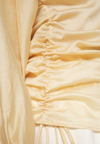 Satiinikangas yläosa pehmeässä beigessä, jossa on kerätyt yksityiskohdat ja sivuissa nauhat. Sileä rakenne, laskostettu muotoilu ja mukava istuvuus.
