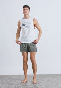 Biały bezrękawnik z czarnym logo "Emporio Armani", zestawiony z szarymi, pasiastymi kąpielówkami. Model stoi boso na fakturowanej powierzchni.