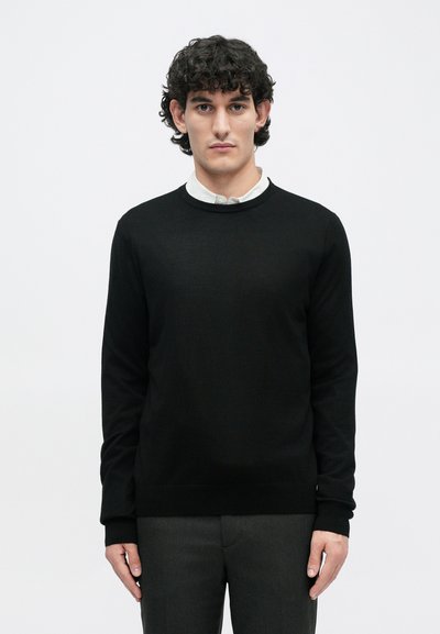 Pull noir avec un col rond, des poignets et un ourlet côtelés. Porté par-dessus une chemise blanche à col, présentant une texture lisse et un design classique.