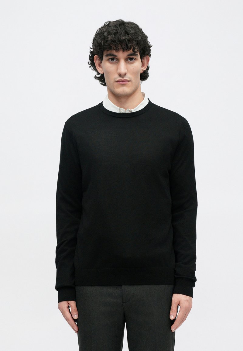 Sort sweater med rund halsudskæring, ribbede manchetter og søm. Bæret over en hvid skjorte med krave, der har en glat tekstur og klassisk design.