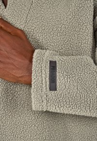 Szürke plüss dzseki texturált felülettel, a mandzsettán a "PEGADOR" feliratú címke található. Közelkép a kézről, ami az anyagon van.