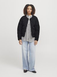Veste bomber noir matelassée avec col rond et fermeture à boutons, portée sur une chemise rayée blanche et noire, associée à un jean large bleu clair.