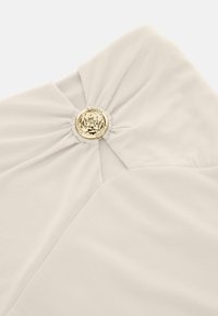 Tessuto a coste beige con una torsione centrale e un accento di logo circolare dorato, creando un design testurizzato e distintivo.
