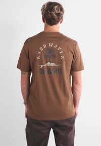 T-shirt en coton marron avec un motif graphique représentant des palmiers et le texte "SAND WAVES" dans le dos. Manches courtes et coupe décontractée.