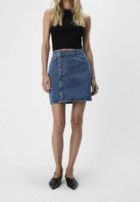 Minifalda de mezclilla en azul, con cierre de botones en el frente, bolsillos delanteros y corte recto. Lleva puesta una camiseta corta negra de canalé y zapatos negros.